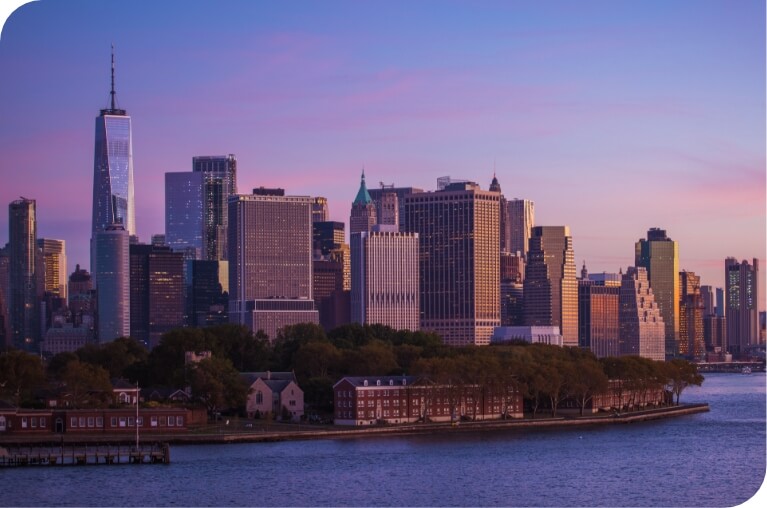 A scenic view of a city skyline at sunset, featuring tall skyscrapers including the One World Trade Center, with a waterfront and small buildings in the foreground.