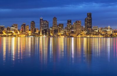 A panoramic photo of the Seattle skyline at night. The Space Needle is visible on the left, and the city lights reflect in the water below. The sky is a deep blue with some clouds.