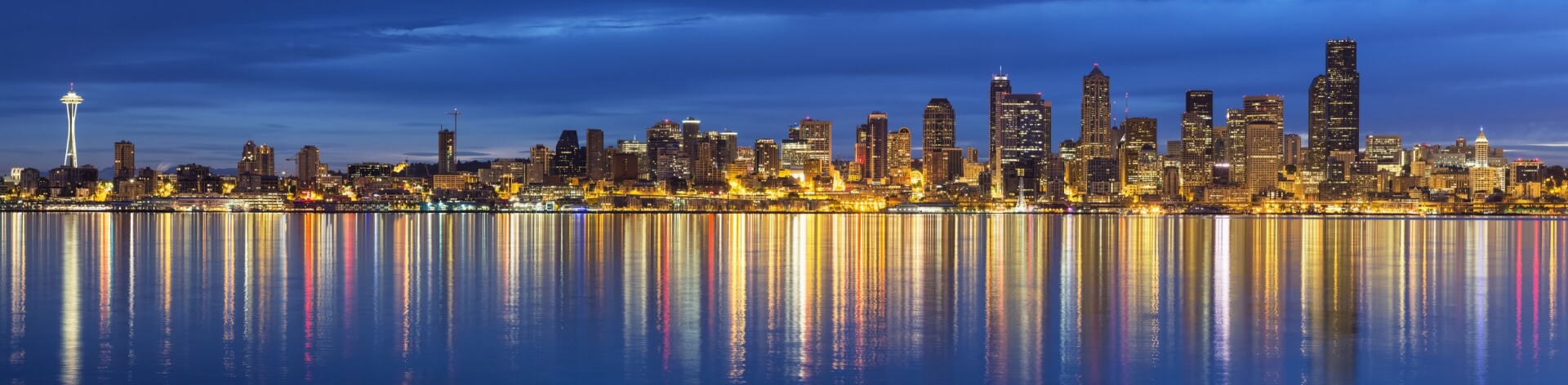 A panoramic photo of the Seattle skyline at night. The Space Needle is visible on the left, and the city lights reflect in the water below. The sky is a deep blue with some clouds.