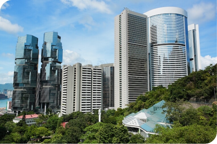 A cityscape with several tall, modern skyscrapers against a blue sky with white clouds. Lush green hills are visible in the foreground.